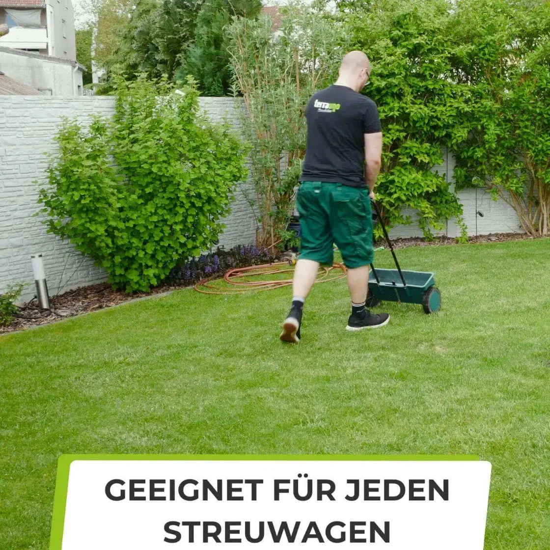Ein Mann in einem schwarzen Hemd und grünen Shorts benutzt einen grünen Streuer für die Rasenpflege auf einem gepflegten Rasen. Im Hintergrund sind Büsche, Bäume und ein weißer Zaun zu sehen. Text: TerraUno Rasendünger - Sparabo: GEEIGNET FÜR JEDEN STREUWAGEN.